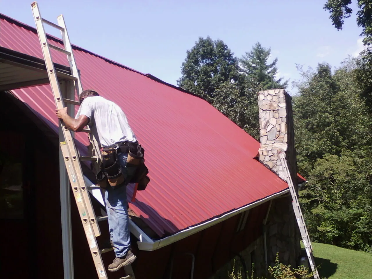 Roofer on a ladder beside a red metal roof and stone chimney — Soffit & Fascia Repair by Roofing Brothers in Saunderstown