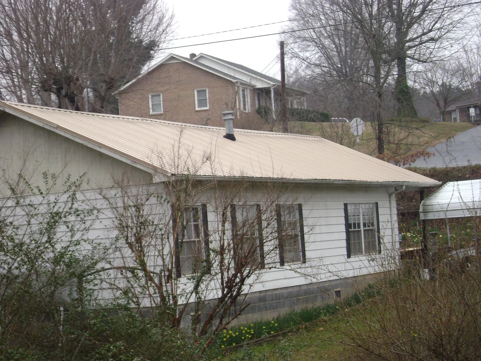 Tan metal roof on a white sided house set into a hillside — Commercial Roofing by Roofing Brothers in Saunderstown